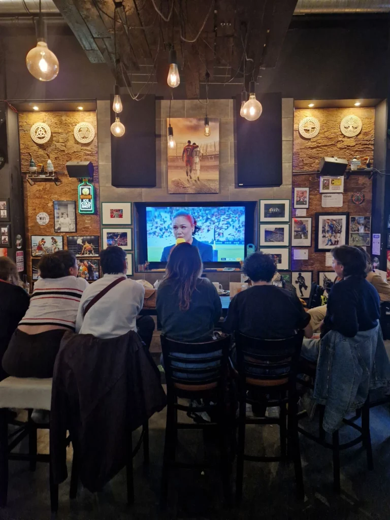 5 femmes de dos regardant un match de sport féminin sur grand écran dans un bar